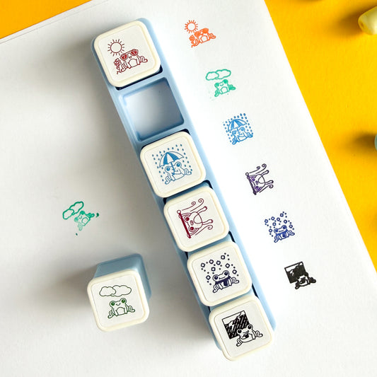 Display of what the frog stamps look like used. The designs: Orange Sunny Day Frog, Green Cloudy Day Frog, Blue Rainy Day Frog, Magenta Windy Day Frog, Blue Snowy Day Frog, Black Stormy Day Frog. The stamps are in their blue holder with the Cloudy Day one out and to the left. There is an example of each design used to the right of the container. The Cloudy Day one has an additional sample on the left. The stamps are shown on white printer paper and there is yellow paper in the top right hand corner.