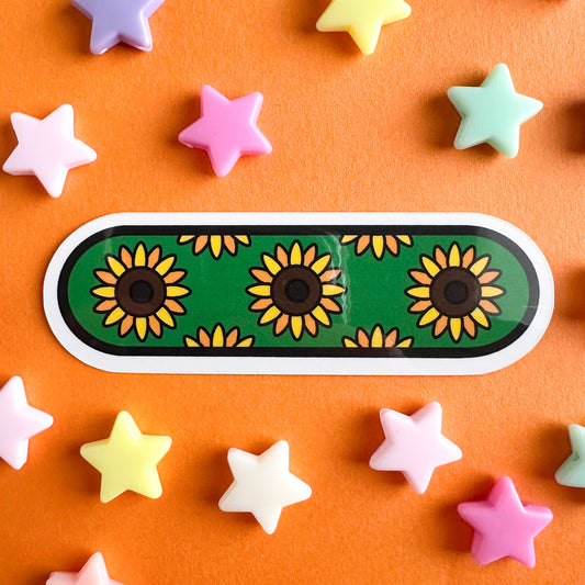 A capsule shaped sticker with illustrations of sunflowers on a green background. The sticker is displayed on an orange piece of paper with plastic star beads around it.