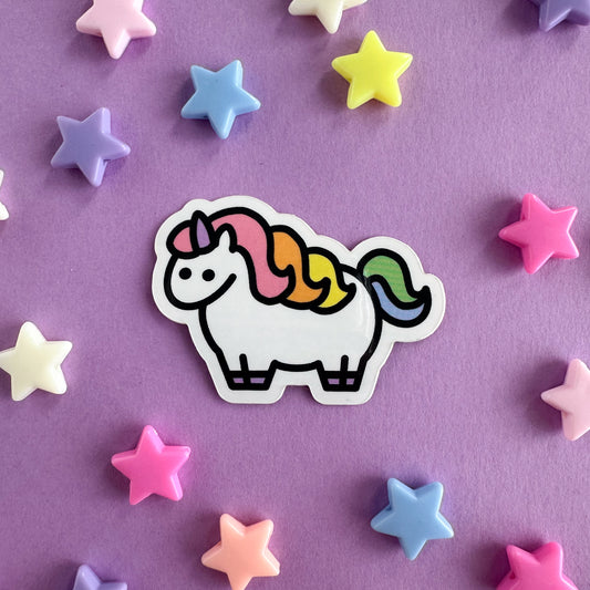 A cute chubby unicorn vinyl sticker in a pastel rainbow color scheme. The sticker is on a lavender paper background with pastel star pony beads strewn around it. 