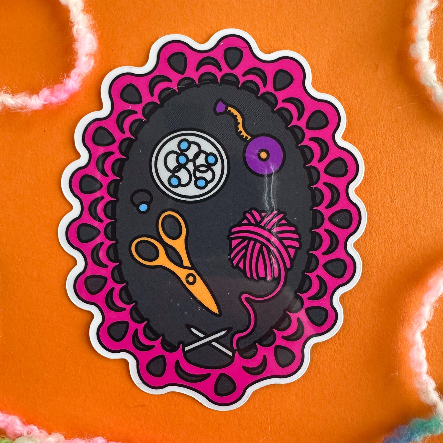 A vinyl sticker featuring an illustration of a knitting project on the needles with various knitting tools like scissors and a tape measure around the knitting. The sticker is on an orange paper background with pastel yarn strewn around it. 