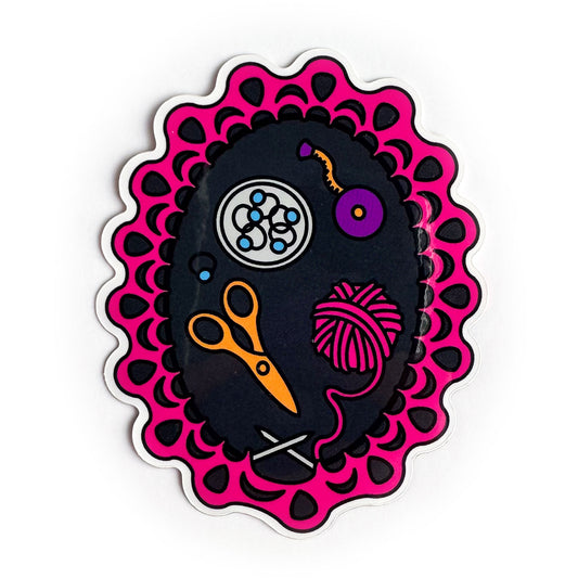 A vinyl sticker depicting a knitting project still on the needles with knitting tools inside of it including scissors, stitch markers, a pink yarn ball and a tape measure. 