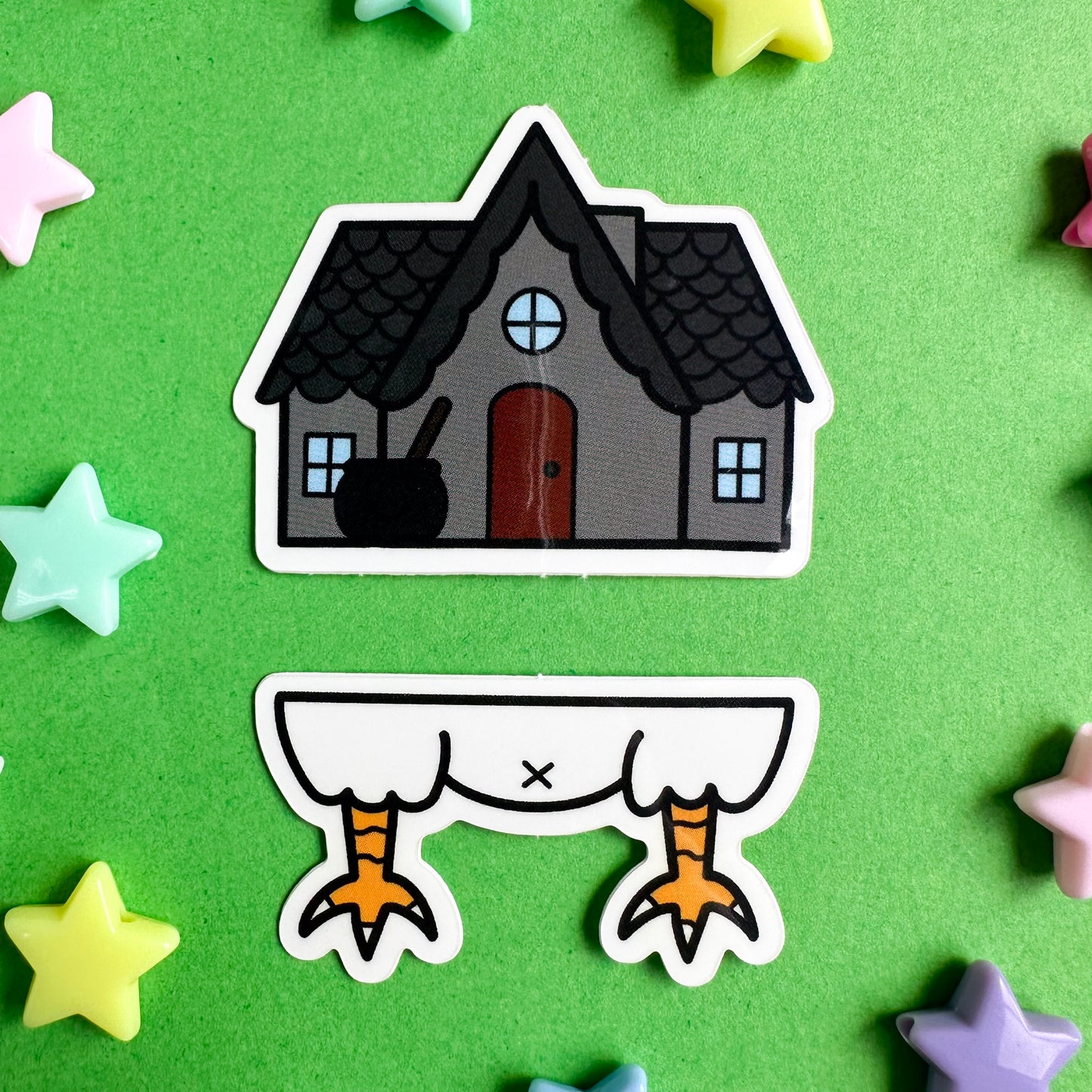 A sticker set that comes together to be Baba Yaga's house, the top sticker is a grey house with a peaked roof and a cauldron outside. The bottom sticker is a chicken butt. The stickers are sitting on a green background with plastic star beads around them. 