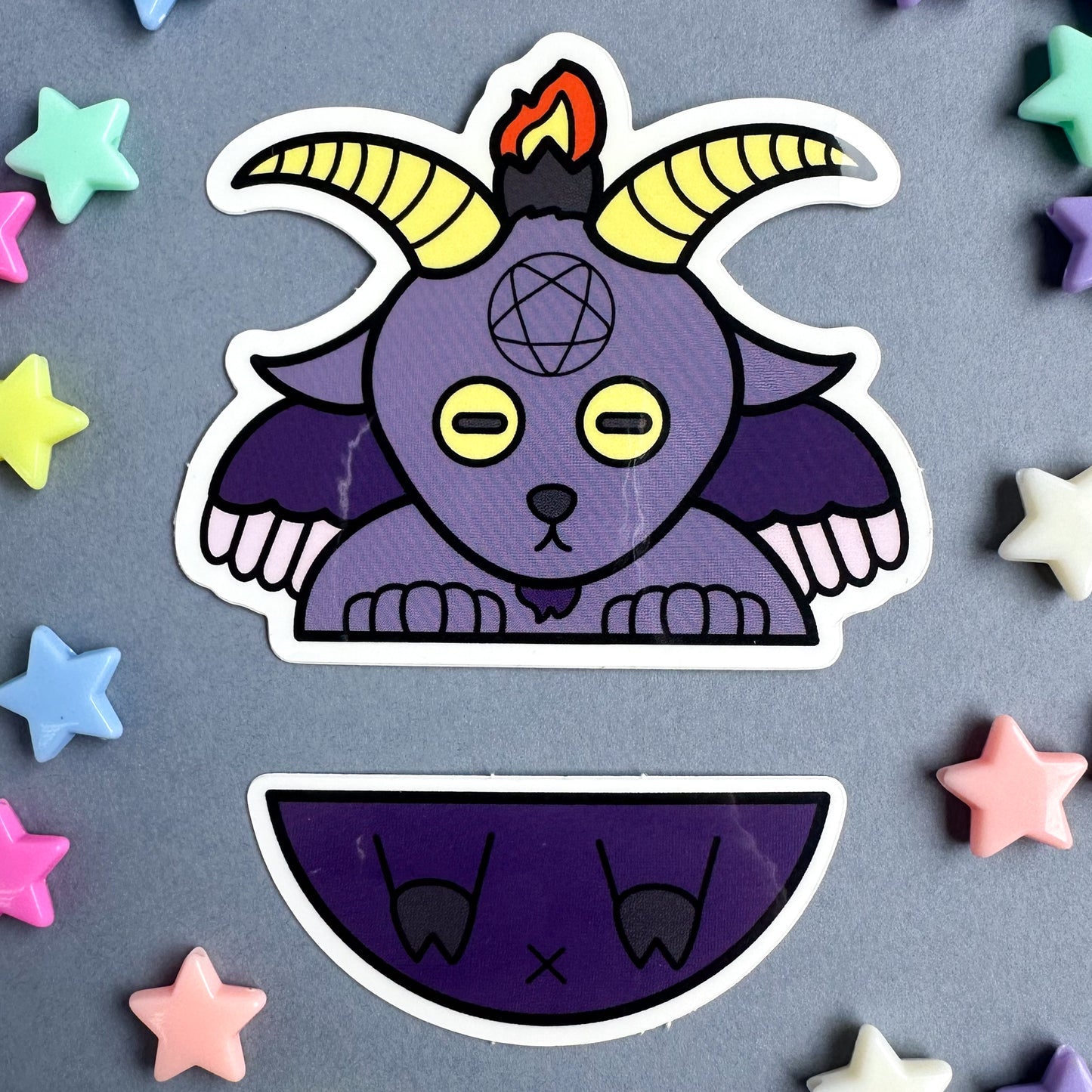 Two stickers that come together to form a cute illustration of Baphomet. The stickers are sitting on a grey background with pastel star beads around it.