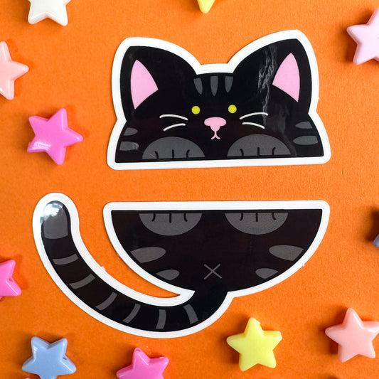 Two stickers that come together to form a black cat that has been split in half horizontally. The stickers are sitting on an orange background with plastic pastel star beads around them.