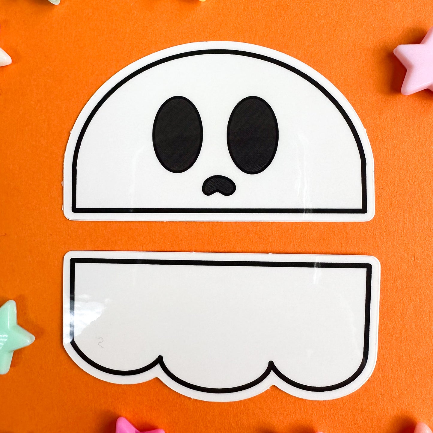 A sticker set of a ghost that has been cut in half horizontally, the top half is the head and the bottom part is the swoop bottom. The stickers are on an orange background star beads around them. 