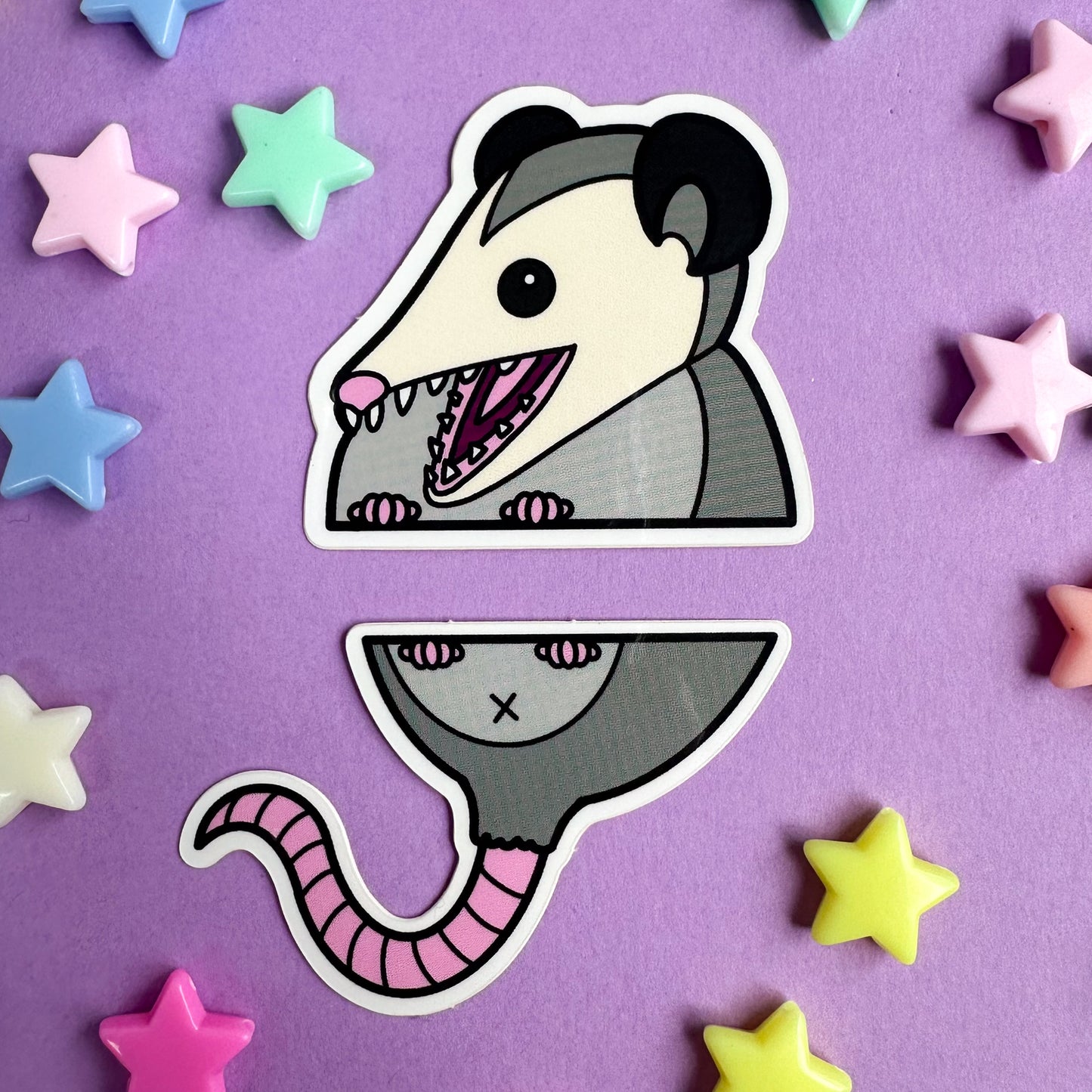 A pair of stickers that come together to form the top and bottom half of a cute opossum. The stickers are on a purple background with pastel stars around it.