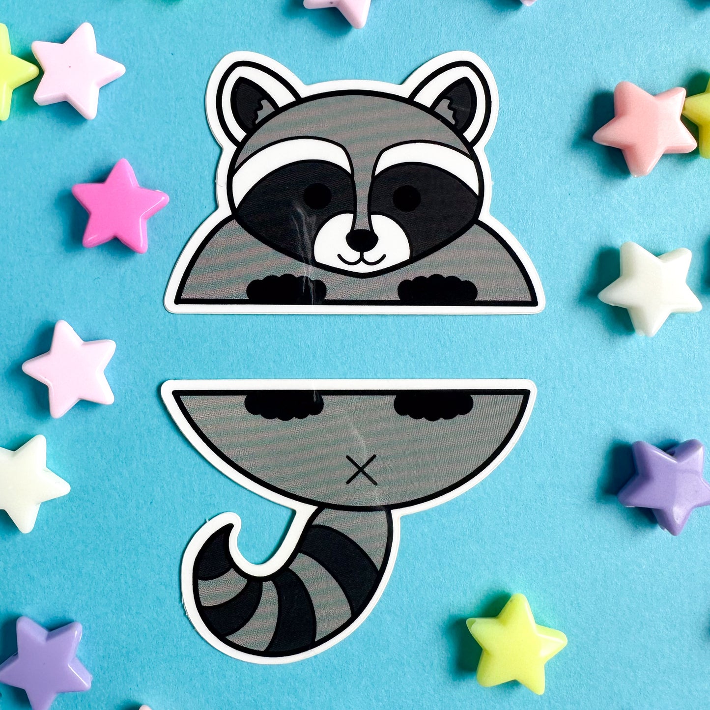 Two stickers that come together to form a cartoony illustration of a raccoon. The stickers are on a blue paper background with star beads scattered around them. 