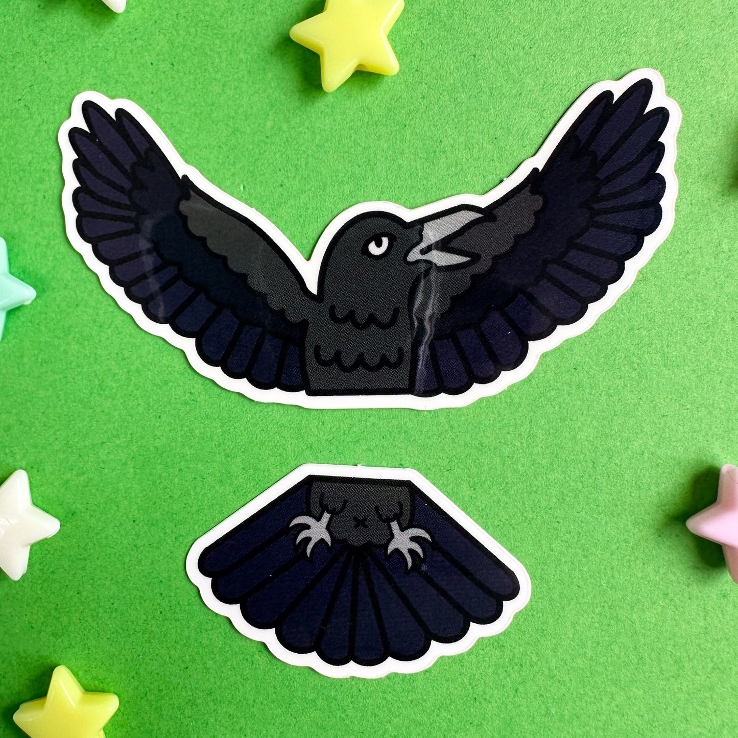 Two vinyl stickers that form a raven cut in half horizontally. The stickers are on a green paper background with star beads around them. 