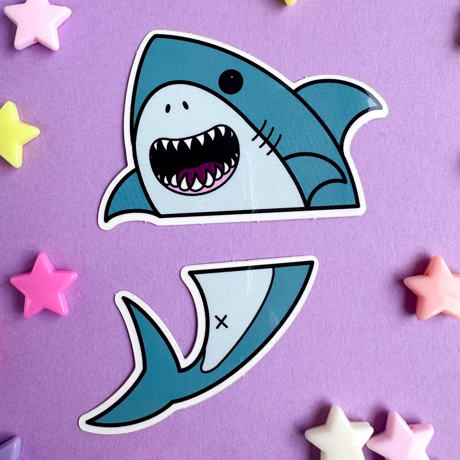 Two vinyl stickers that come together to form a cute illustration of a shark that is divided horizontally. The stickers are on a purple paper background scattered with plastic star beads.