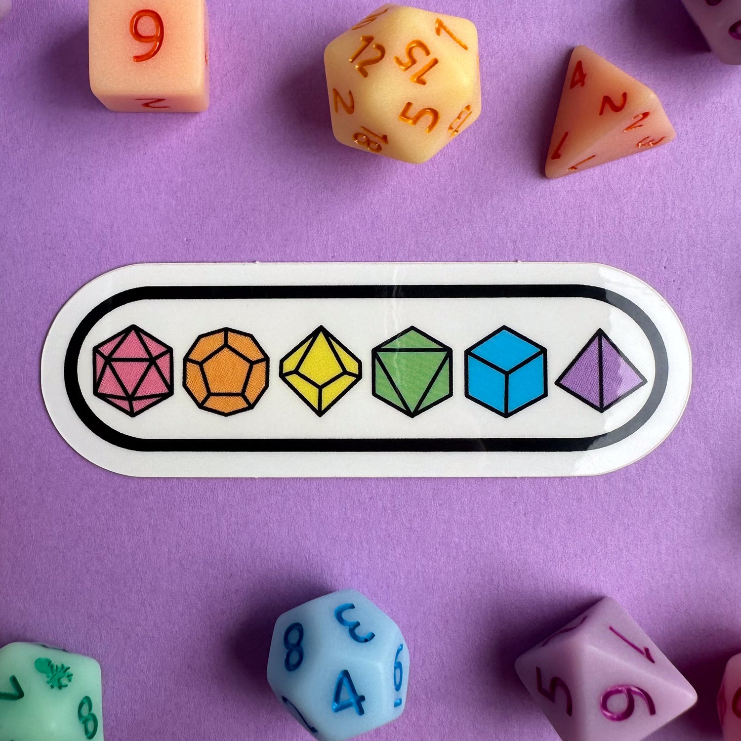 An oval shaped sticker with pastel rainbow dice on it.The sticker is on a purple background with matching real polyhedral pastel dice around it. 