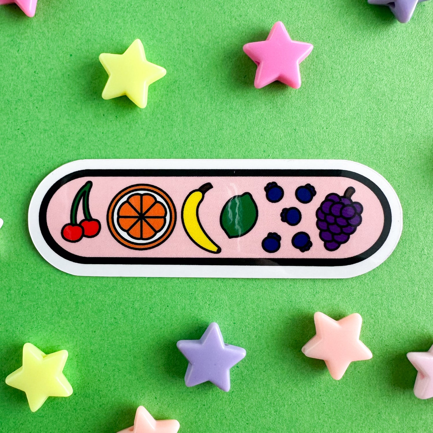 A capsule shaped sticker with illustrations of fruit on it including cherries, an orange, a banana, a lime, blueberries and grapes. The background of the sticker is light pink. The sticker is on a green paper background with pastel plastic star beads around it. 