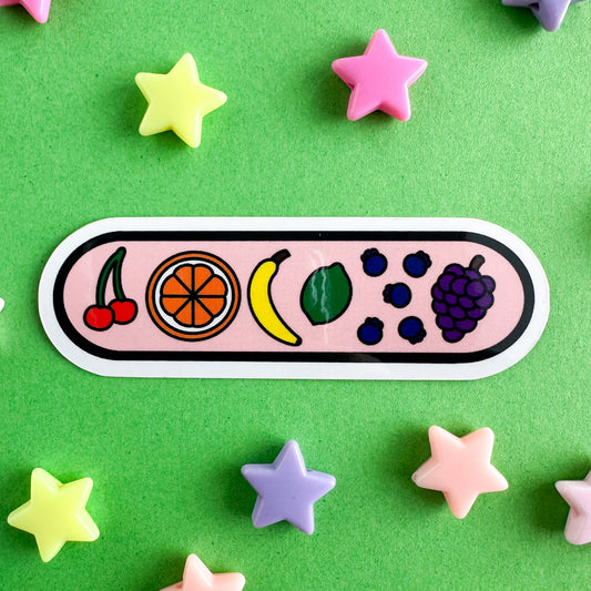 A capsule shaped sticker with illustrations of fruit on it including cherries, an orange, a banana, a lime, blueberries and grapes. The background of the sticker is light pink. The sticker is on a green paper background with pastel plastic star beads around it.