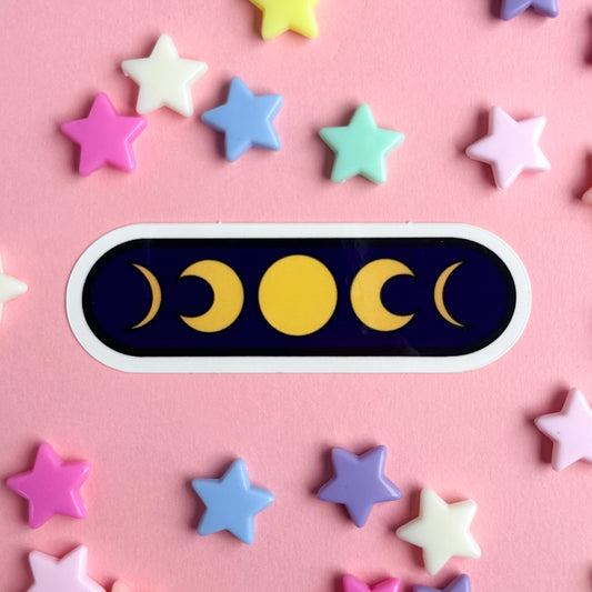A dark oval sticker with yellow illustrations of phases of the moon on it. The sticker is on a pink paper background with pastel stars around it.