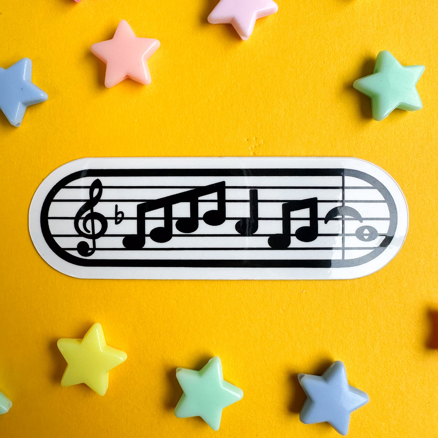 A capsule shaped sticker with a music staff and notation on it. The sticker is on a yellow piece of paper with pastel rainbow star beads around it.  