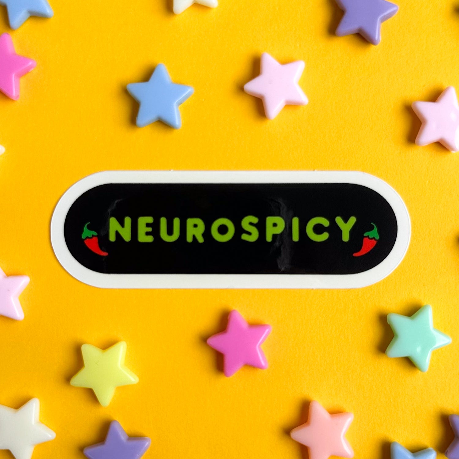 The word neurospicy on an oval shaped sticker, the letters are lime green on a black background framed by chili peppers. The sticker is on a yellow piece of paper with star beads around it. 
