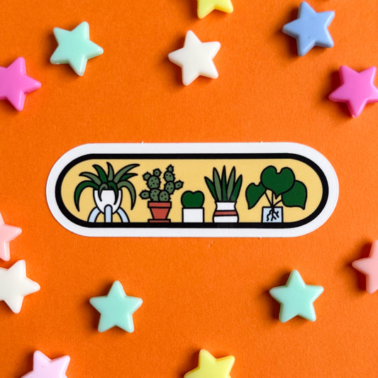 A vinyl capsule sticker with illustrations of houseplants on it including a spider plant, cactus, Hoya and snake plant. The sticker is on an orange background with pastel star beads around it.