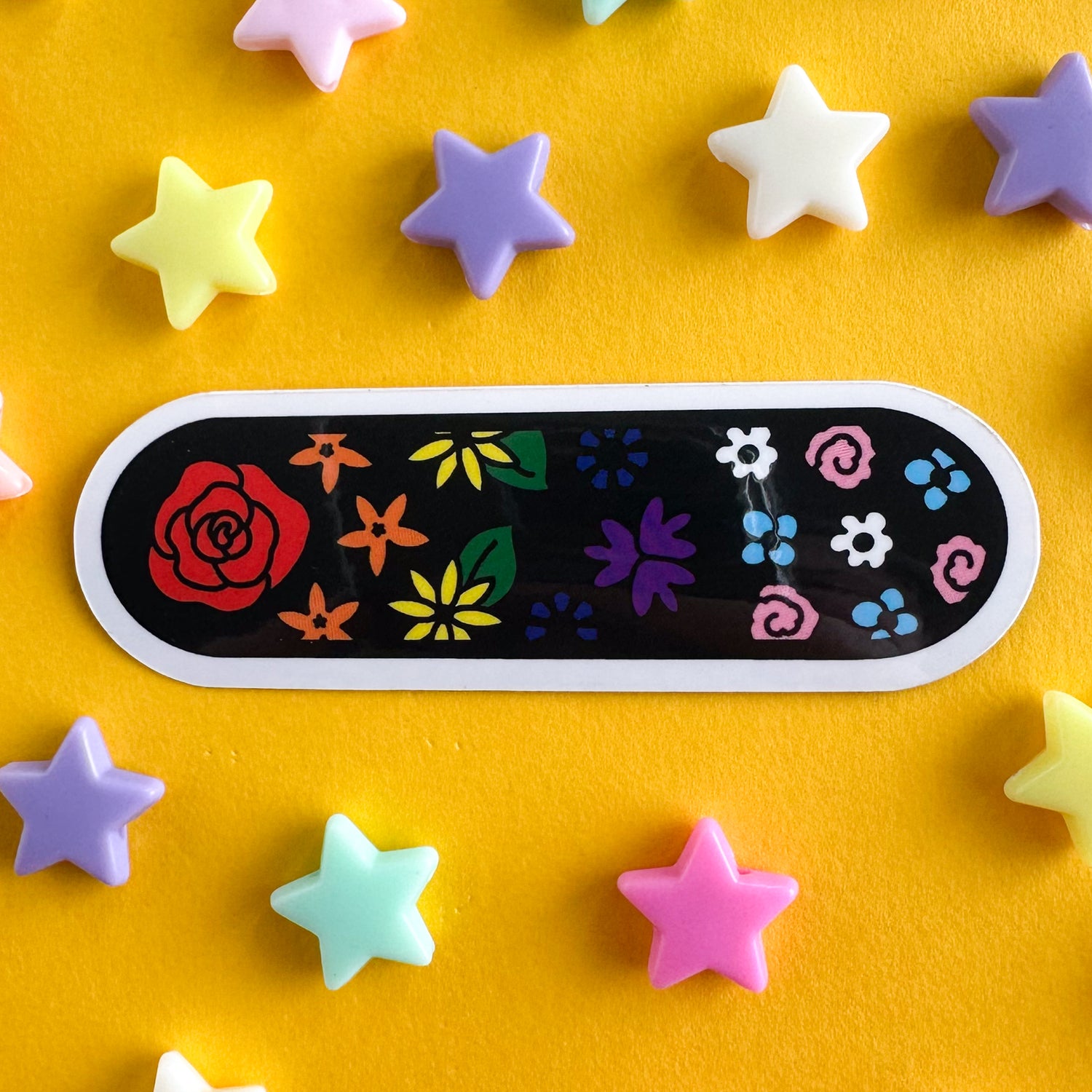 A sticker with a black background and illustrations of flowers on it, the flowers are the colors of the rainbow pride and trans pride flags. The sticker is shaped like a bandaid. The sticker is on a yellow background with pastel plastic pony beads around it. 