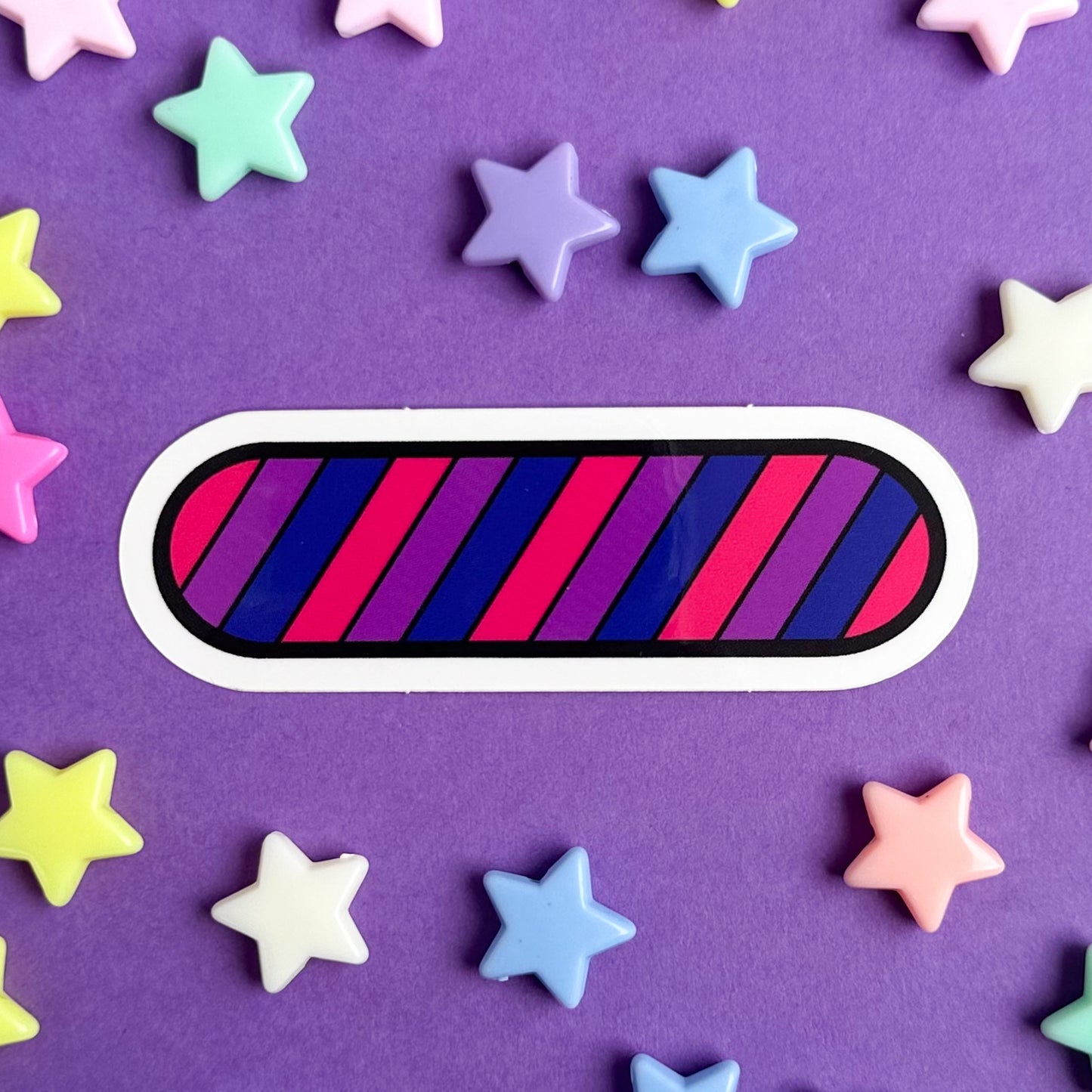 A vinyl sticker in the shape of an oval in the colors of the Bisexual Pride Flag on a purple background with star shaped beads around it. 