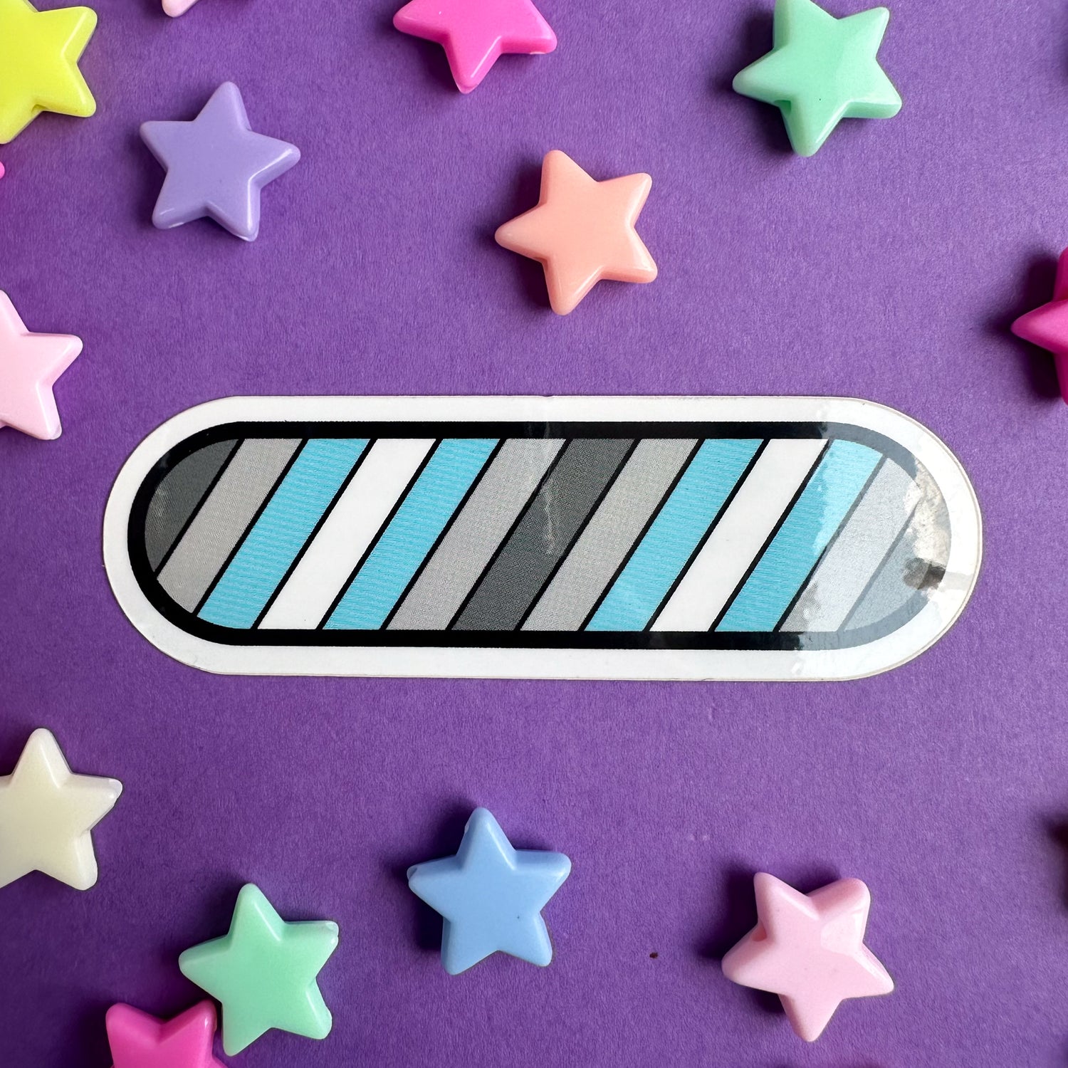 An oval shaped enamel pin with diagonal stripes in the colors of the Demiboy Pride Flag. The sticker is on a purple paper background with plastic star beads around it. 