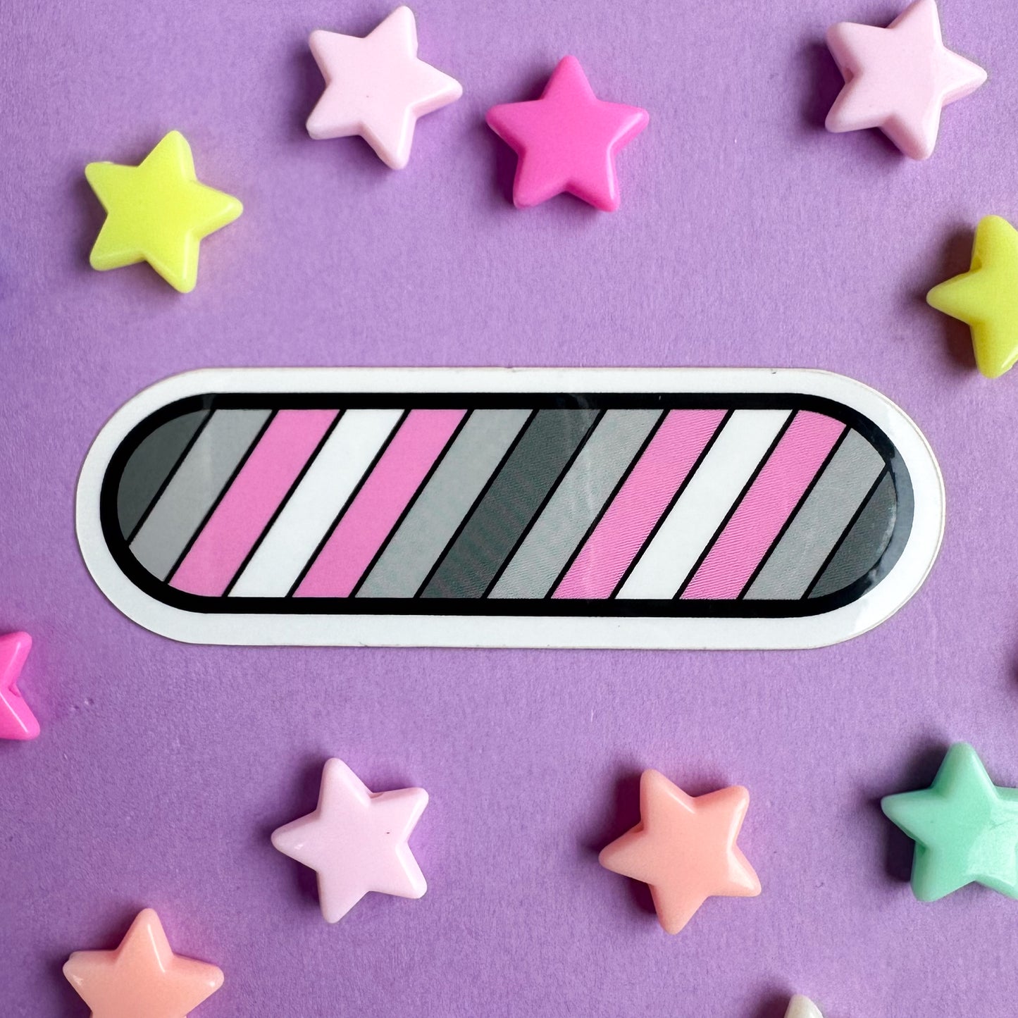 A capsule shaped sticker with stripes in the colors of the Demigirl Pride Flag in diagonal stripes across it.  The sticker is on a purple piece of paper with star beads around it. 