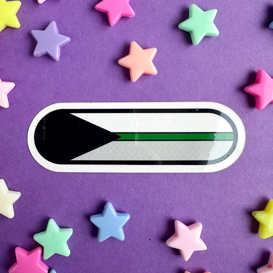 A capsule shaped sticker with the image of the Demiromantic Pride Flag on it. The sticker is on a purple paper background with pastel star beads around it.