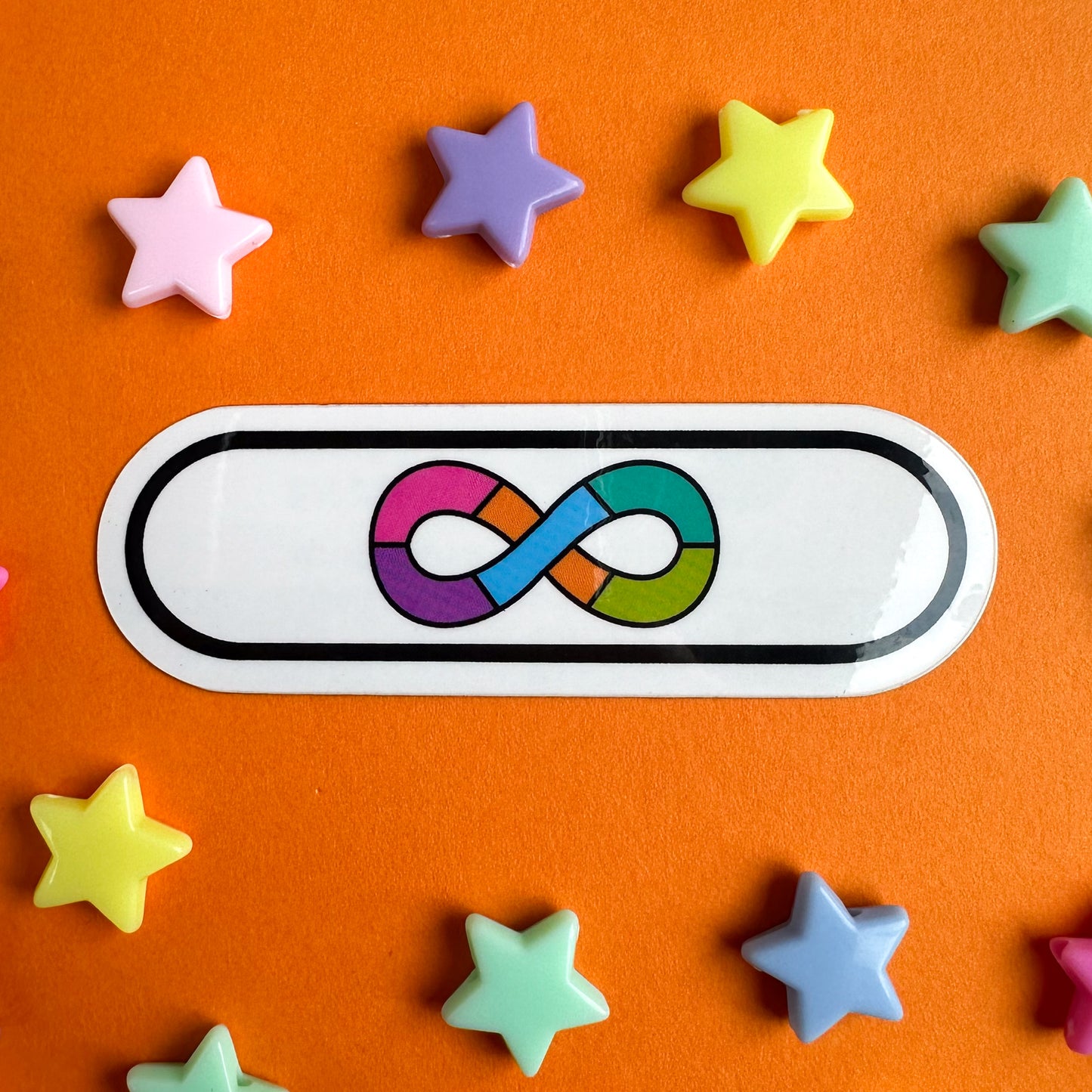 A white vinyl sticker with the rainbow infinity symbol of the Neurodivergent Pride flag. The sticker is on an orange piece of paper with plastic star beads around it. 