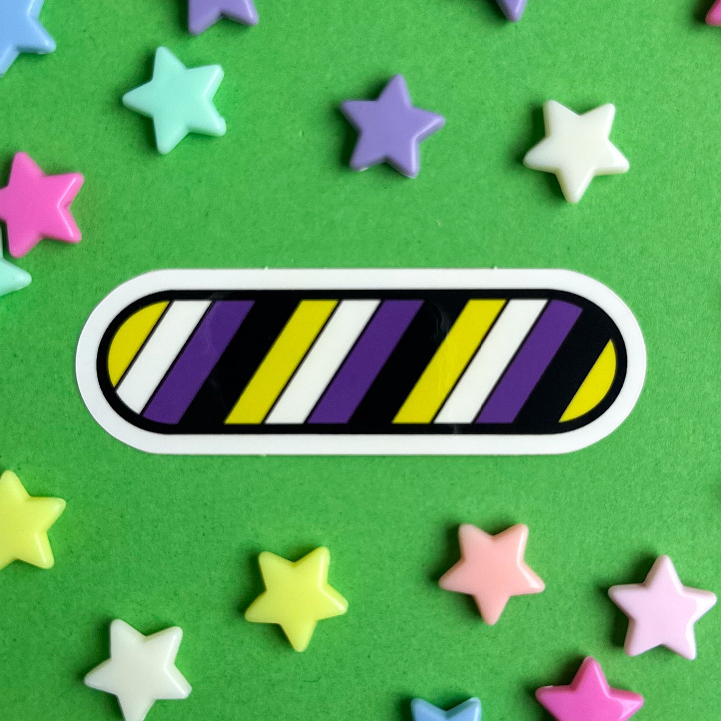 An oval shaped sticker with diagonal stripes of the Nonbinary Pride flag. The sticker is on a green background with pastel star beads around it.