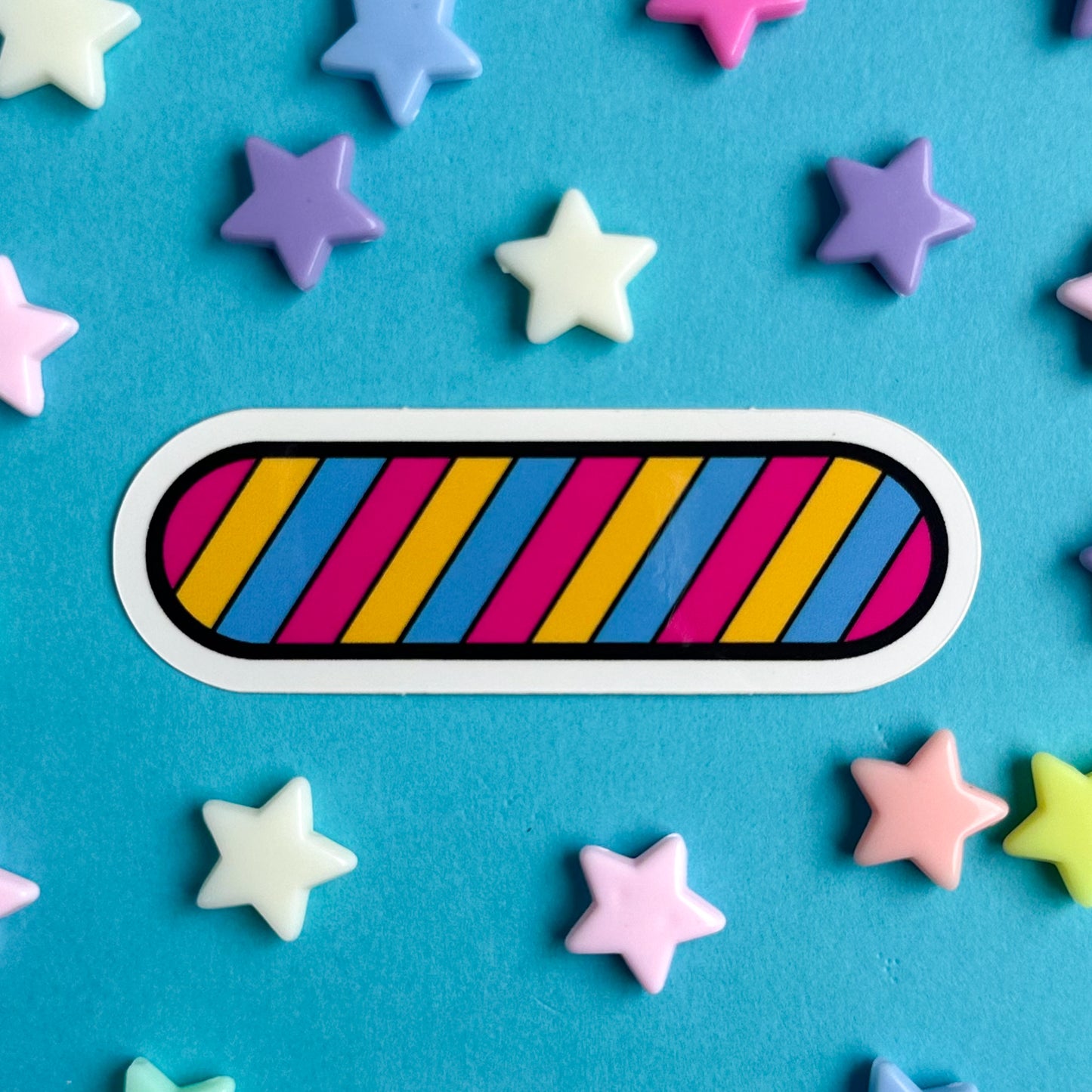 A vinyl sticker shaped like an oval with diagonal stripes of the Pansexual pride flag. The sticker is on a blue background with pastel star beads around it.