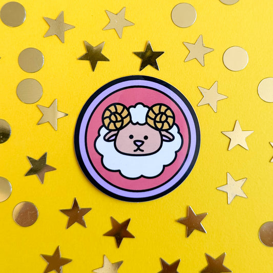 A circular sticker with a cute ram on it to symbolize Aries the sticker is sitting on a yellow background with gold stars and circles around it.