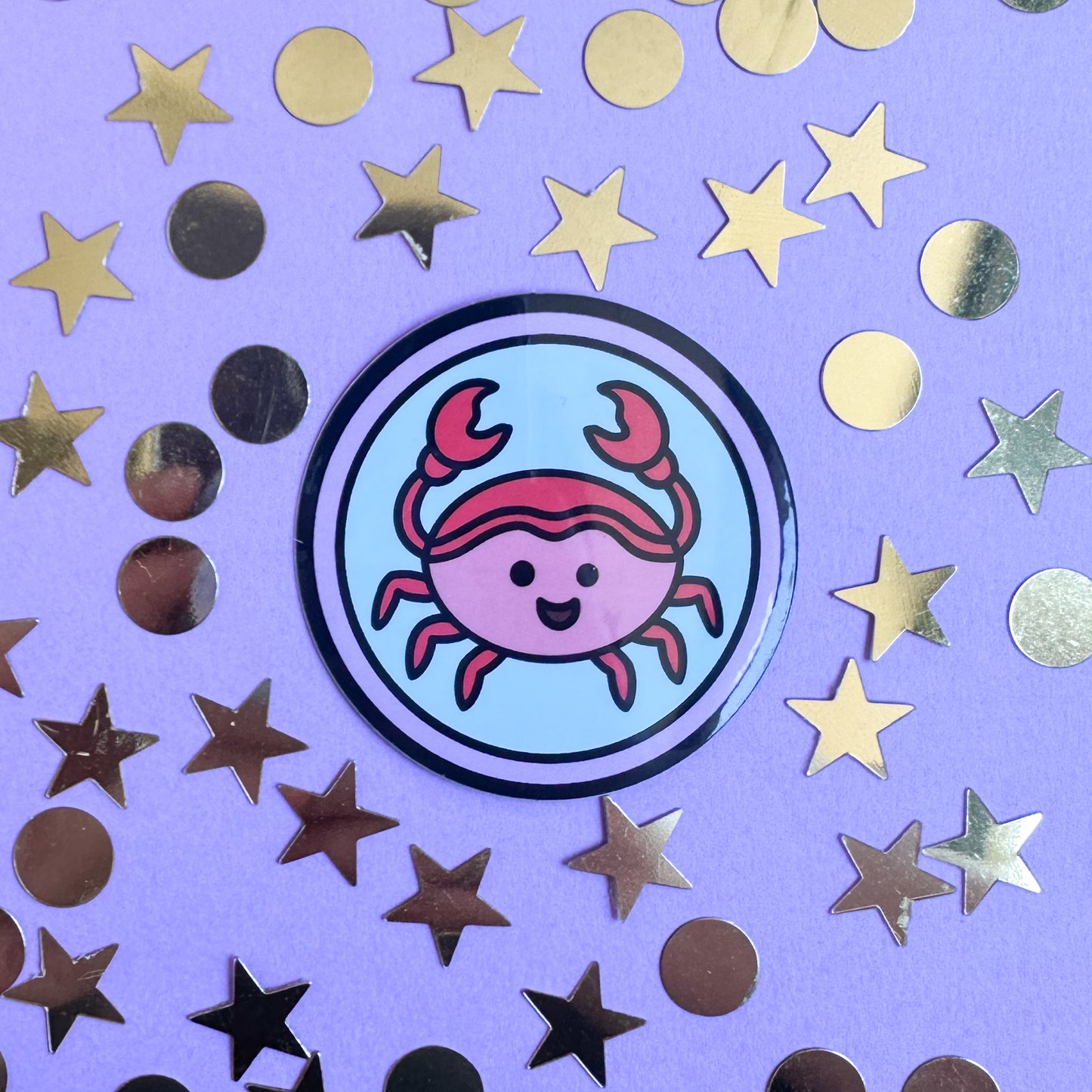 A circle sticker with a cute crab illustration in it to represent the cancer zodiac sign. The sticker is on a purple background with gold glitter stars and circles around it. 
