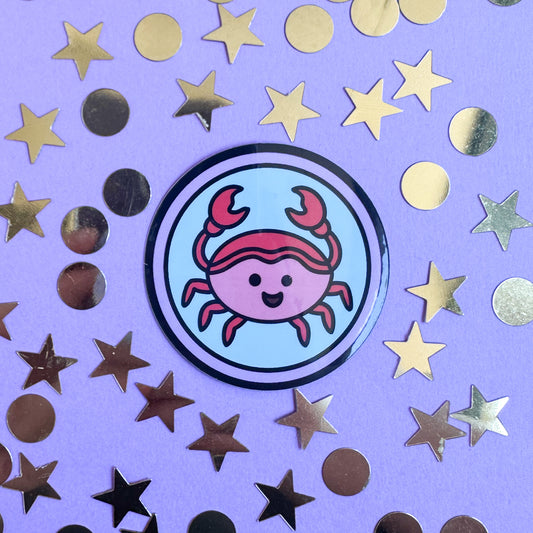 A circle sticker with a cute crab illustration in it to represent the cancer zodiac sign. The sticker is on a purple background with gold glitter stars and circles around it.