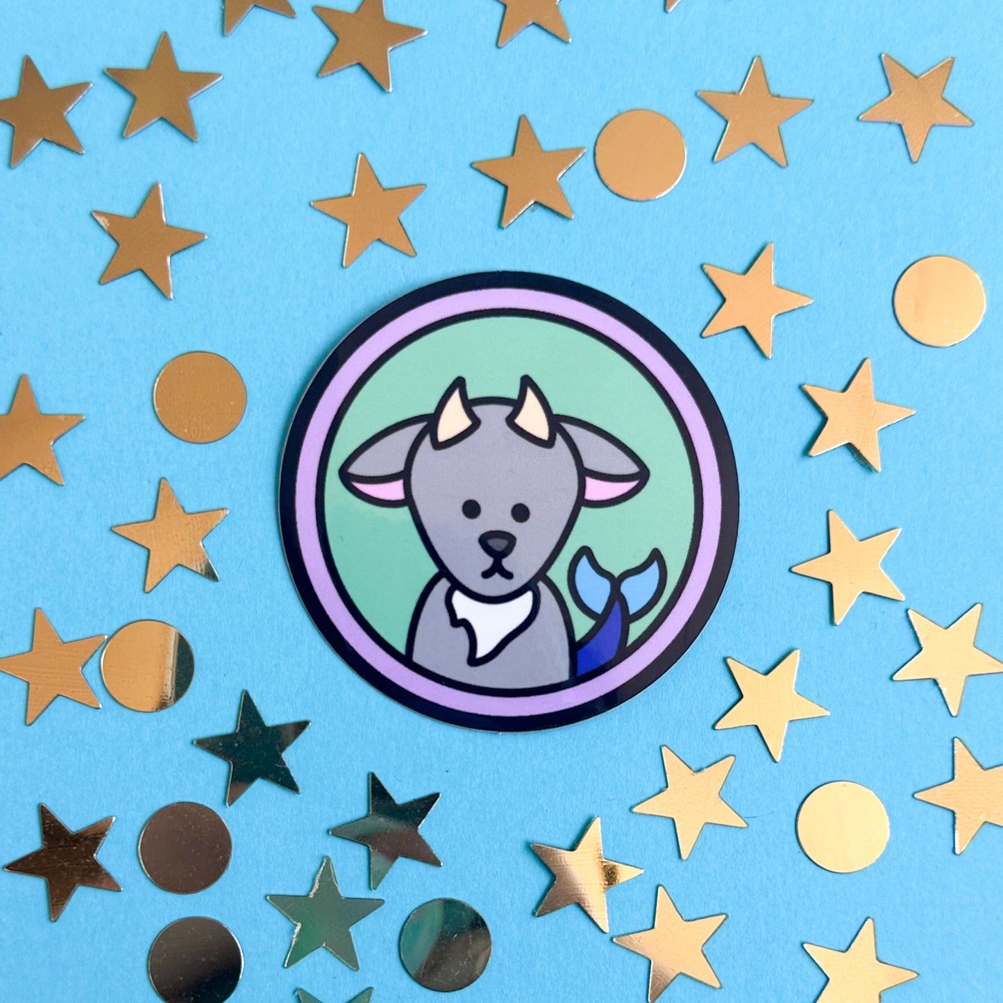 A circle sticker with a cute illustration of a sea goat representing Capricorn as an astrology sign. The sticker is on a blue background surrounded by gold stars and circles. 