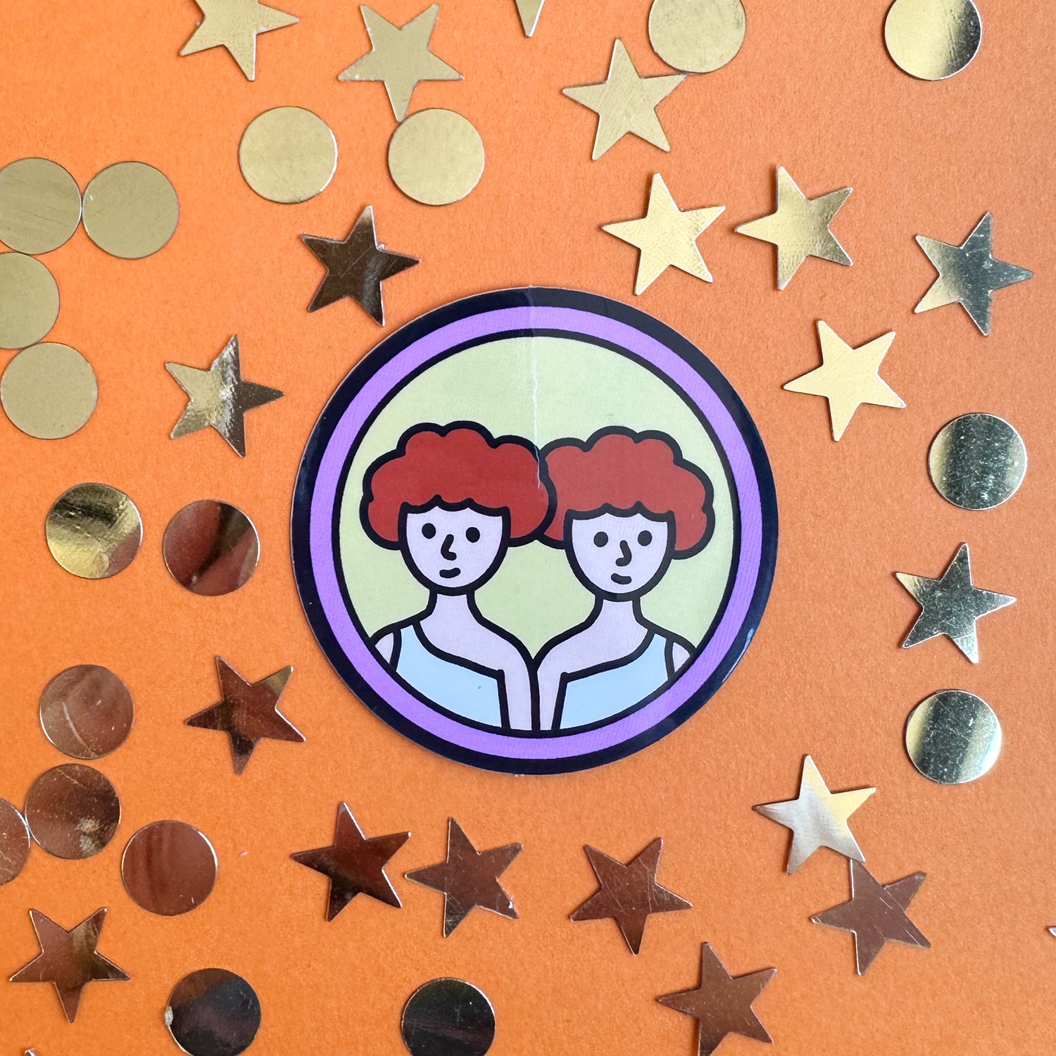 A circular sticker with twins on it with red hair that represent the Gemini zodiac sign. The sticker is on an orange background with gold stars and circles around it. 