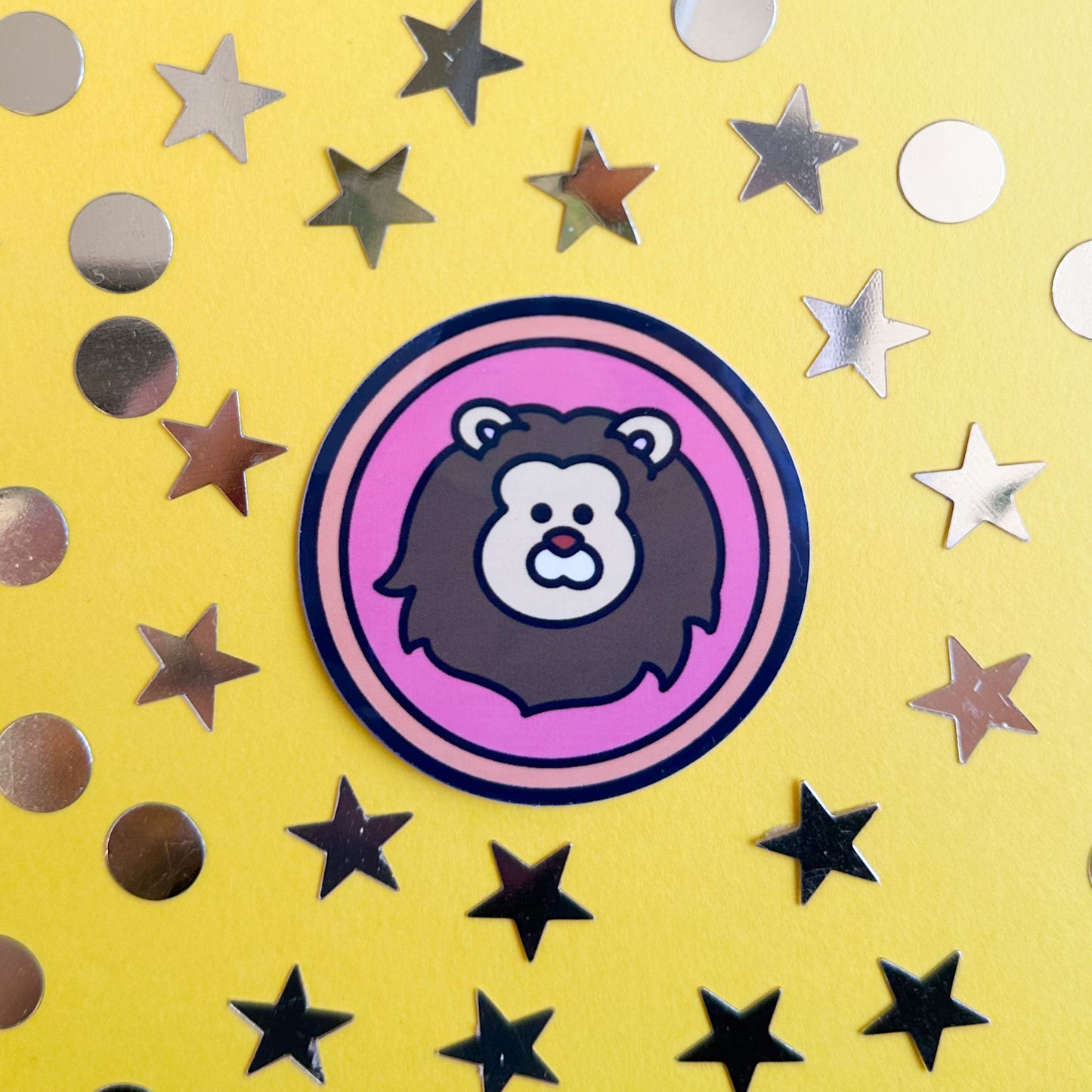 A circle sticker with a cute lion head on it to represent the Leo zodiac sign. The sticker is on a yellow background with gold star and circle confetti around it. 