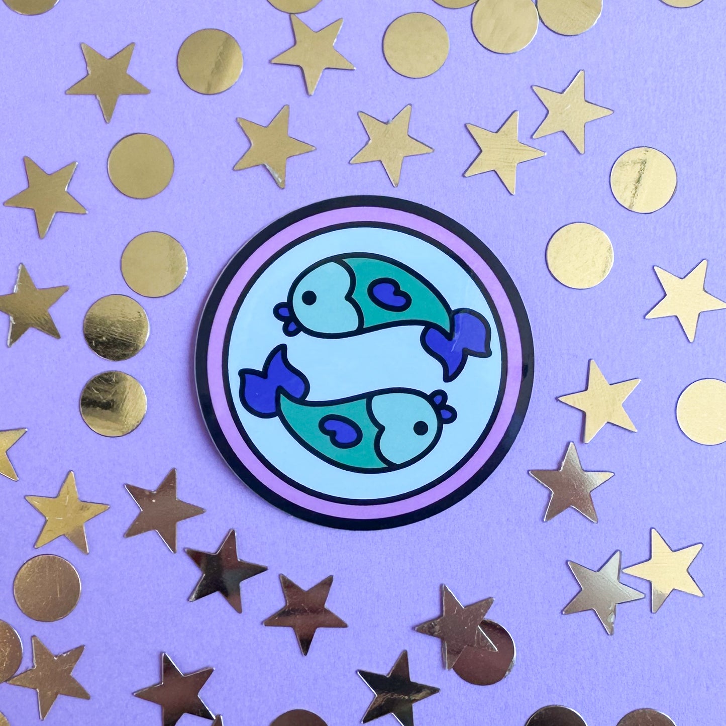 A circle sticker with two fish representing the Pisces zodiac sign. The sticker is on a purple background that has gold confetti in star and circle shapes around it. 