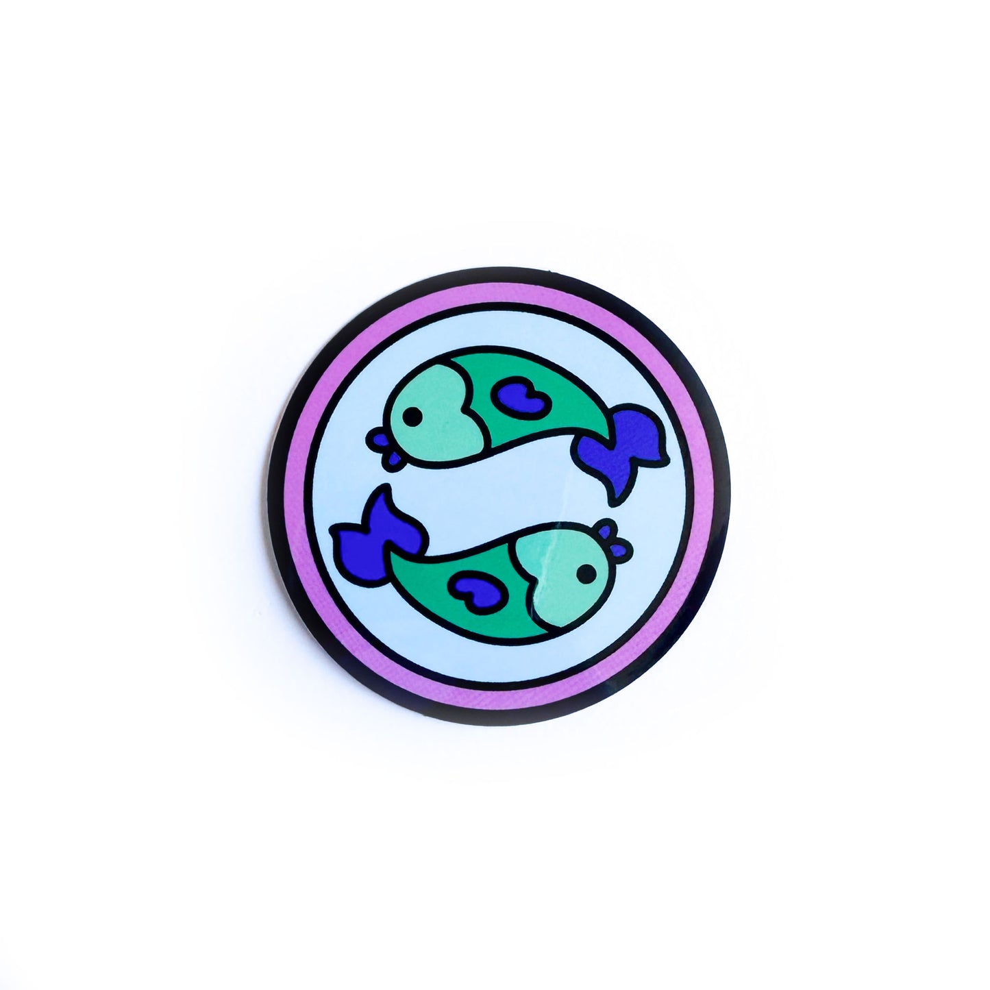 A sticker of a blue circle with a pink background with two green fish swimming in opposite directions on the blue circle to represent the Pisces zodiac sign. 