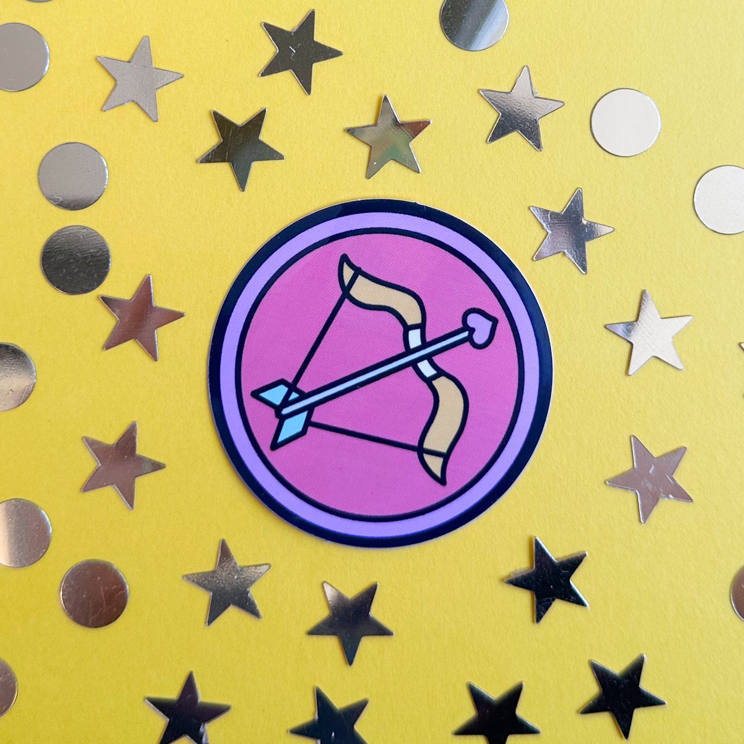 A circle sticker with a cute version of the symbol for Sagittarius in it. The sticker is on a yellow paper background with gold confetti around it. 