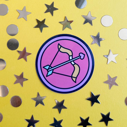 A circle sticker with a cute version of the symbol for Sagittarius in it. The sticker is on a yellow paper background with gold confetti around it.
