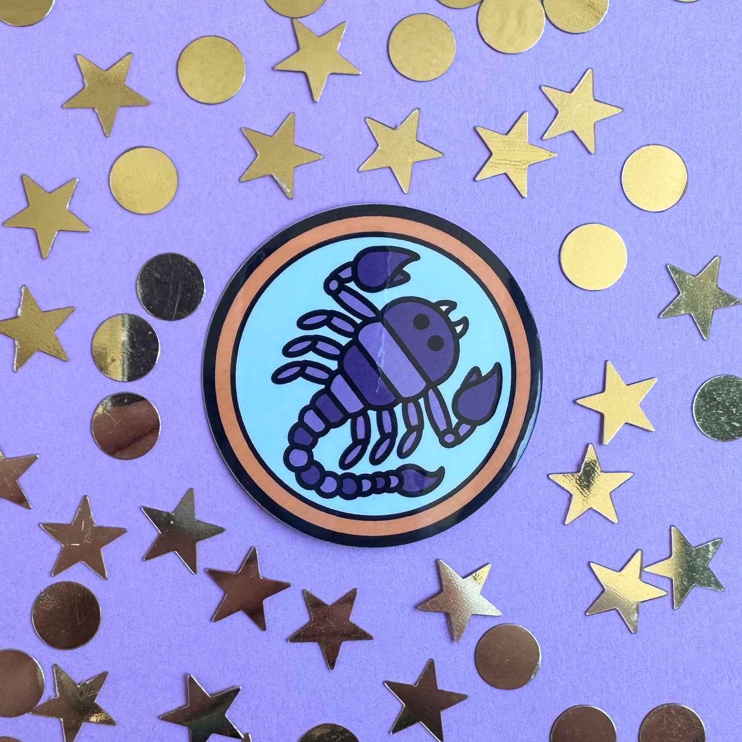 A circle sticker with a purple scorpion in it to symbolize the Scorpio zodiac sign. The sticker is on a purple paper background covered in gold star and circle confetti. 
