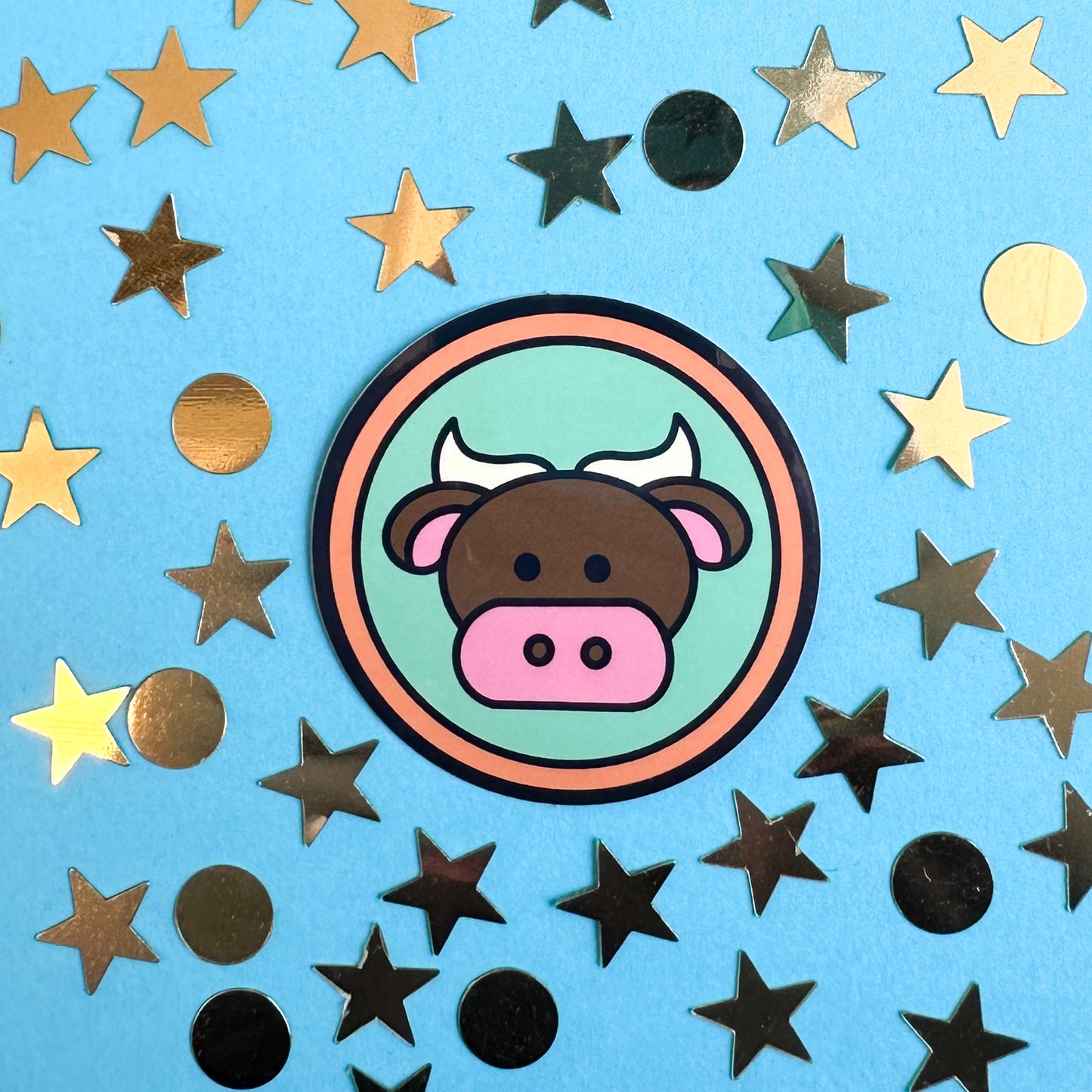 A circle sticker with a bull on it to represent the Taurus zodiac sign. The sticker is on a blue paper background that has gold confetti in star and circle shapes around it. 