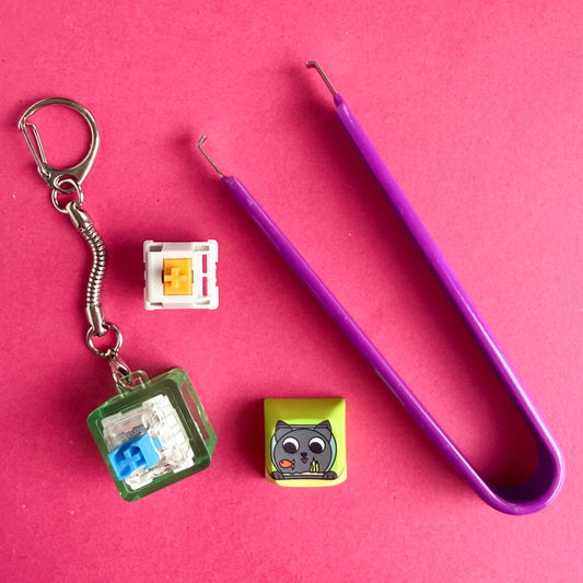 A purple switch puller tool for a mechanical keyboard next to a Clicky Cat keychain and a mechanical keyboard switch. 