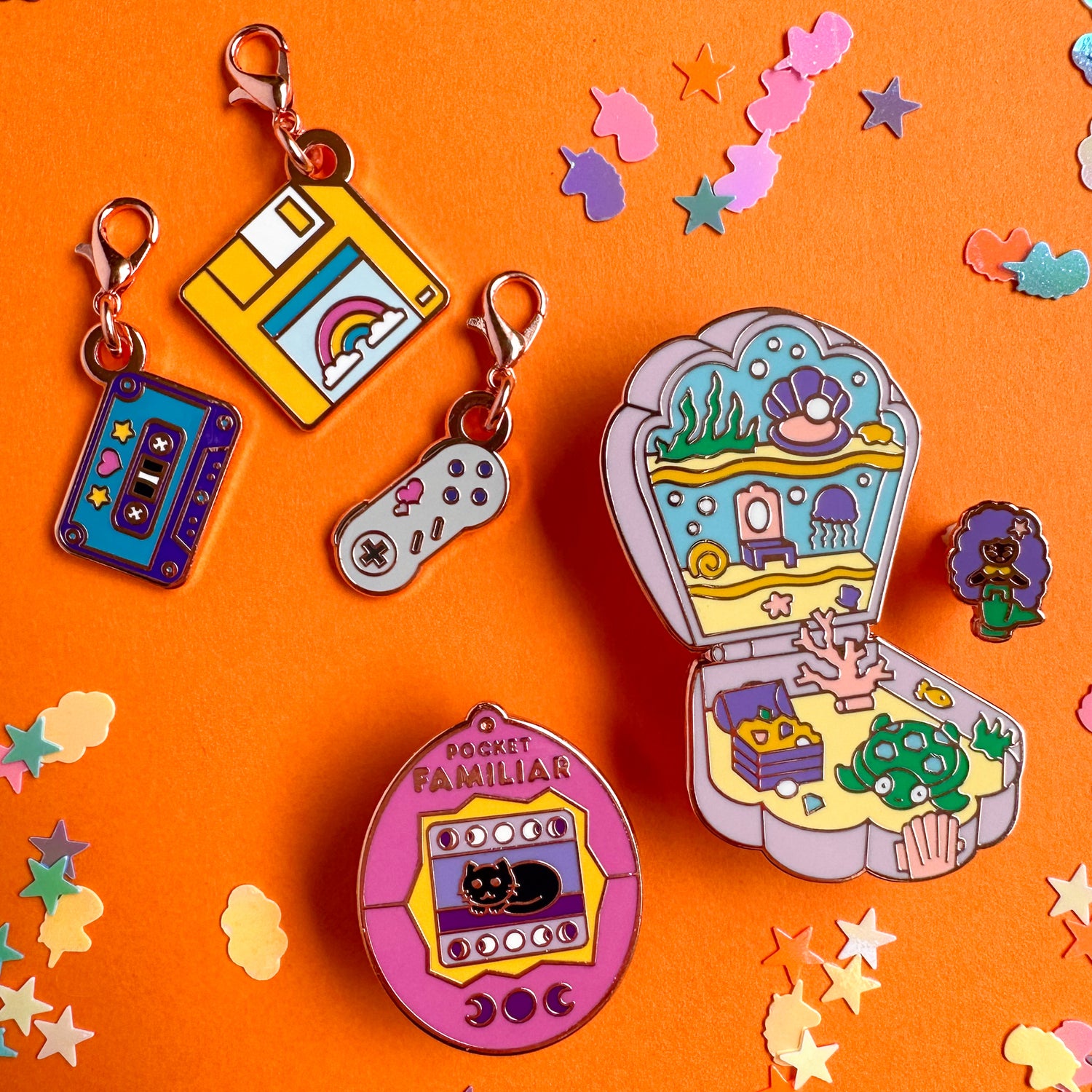 An enamel pin shaped like a vitural pet, a Polly Pocket like mermaid toy set, and 4 charms shaped like a Gameboy, cassette tape, floppy disk, and a Super Nintendo controller all on an orange confetti background.
