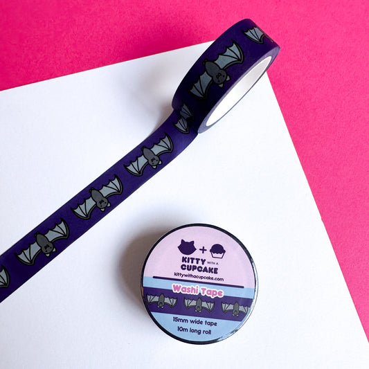 A roll of washi tape with a dark purple background with bats on it stuck to a white piece of paper on a hot pink background. A full roll of packaged washi tape is below.