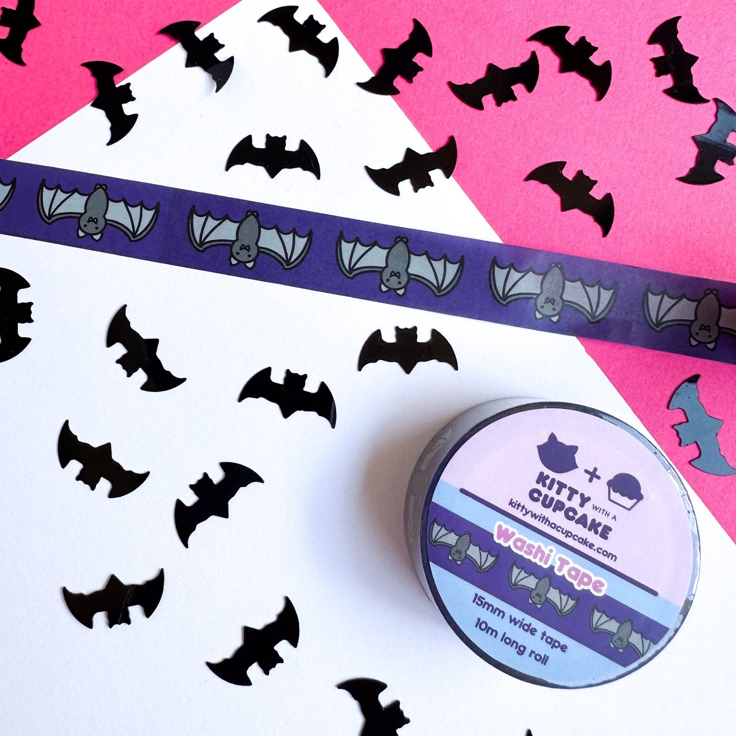 A piece of washi tape stuck to a white piece of paper on a hot pink piece of paper. The tape is dark purple with illustrations of cute bats on it. There is black bat confetti strewn around the tape. A full roll of packaged washi tape is below. 