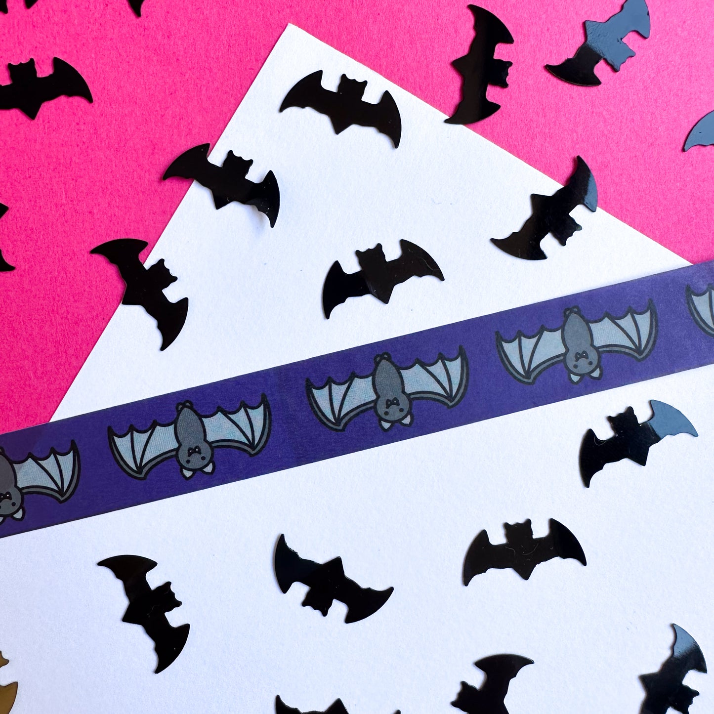 A piece of washi tape stuck to a white piece of paper on a hot pink piece of paper. The tape is dark purple with illustrations of cute bats on it. There is black bat confetti strewn around the tape.