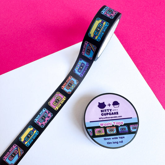 A roll of washi tape stuck to a piece of white paper on top of a hot pink piece of paper. The tape is black with art of colorful cassette tapes on it in a row.