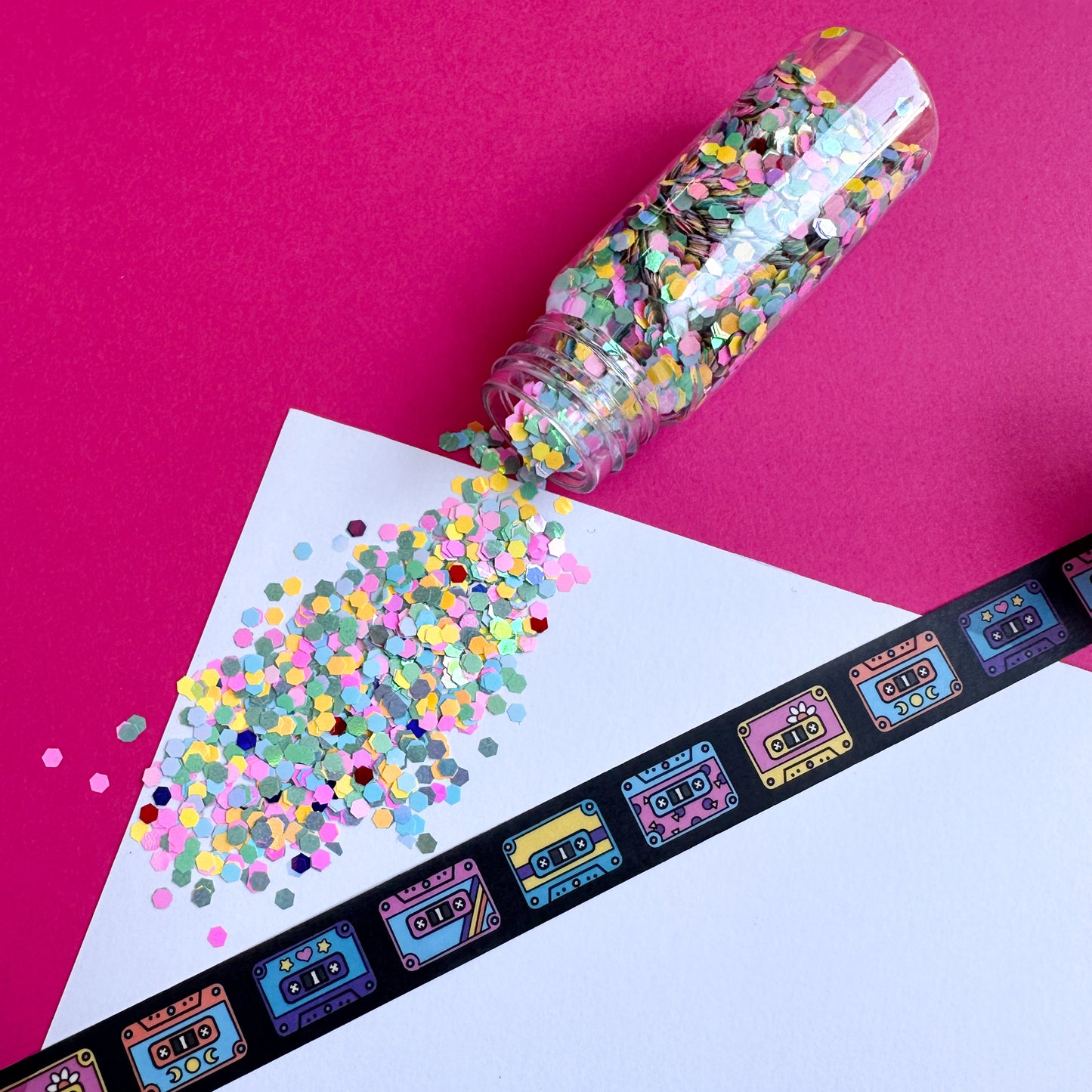 A white piece of paper with spilled pastel confetti coming out of a jar above a piece of washi tape that is black with cute colorful cassette tapes on it. 