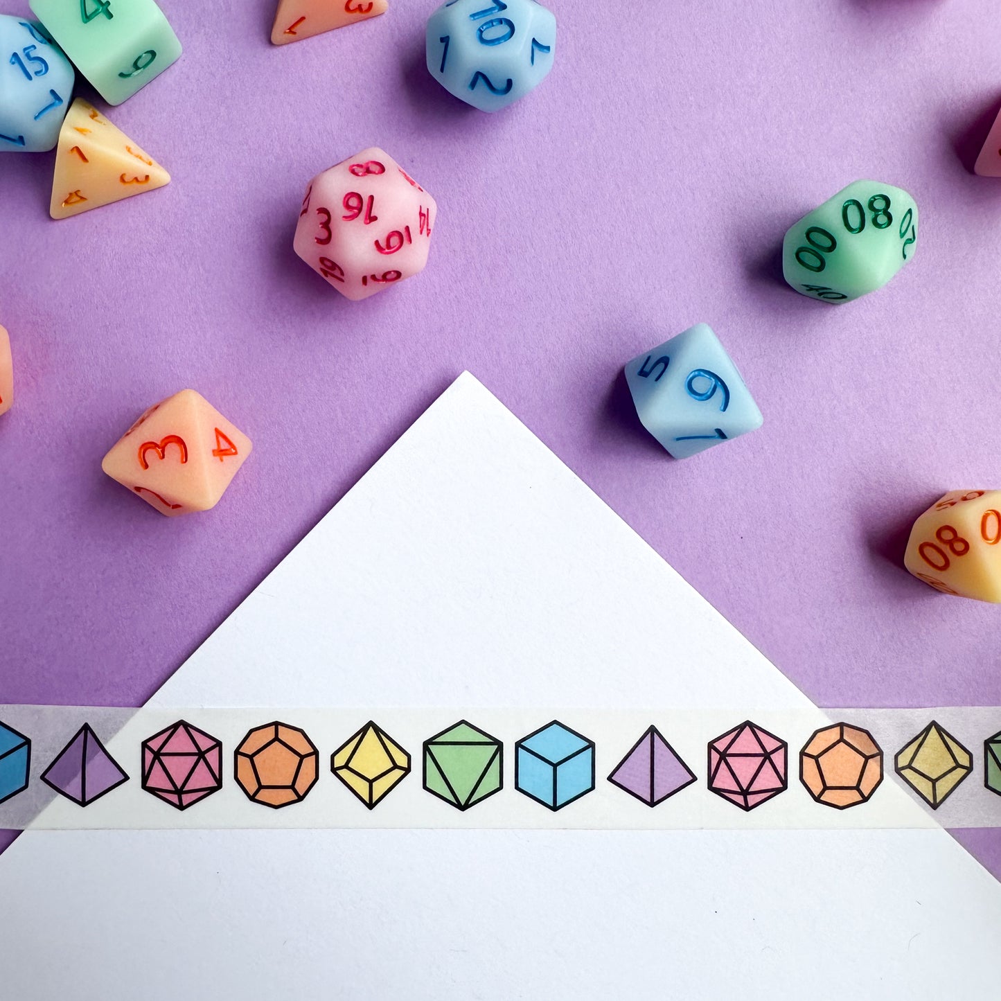 A piece of washi tape with illustrations of DnD dice on it including a D20, D12, D10, D8, D6, and D4. There are real dice that match the colors of the tape strewn around.