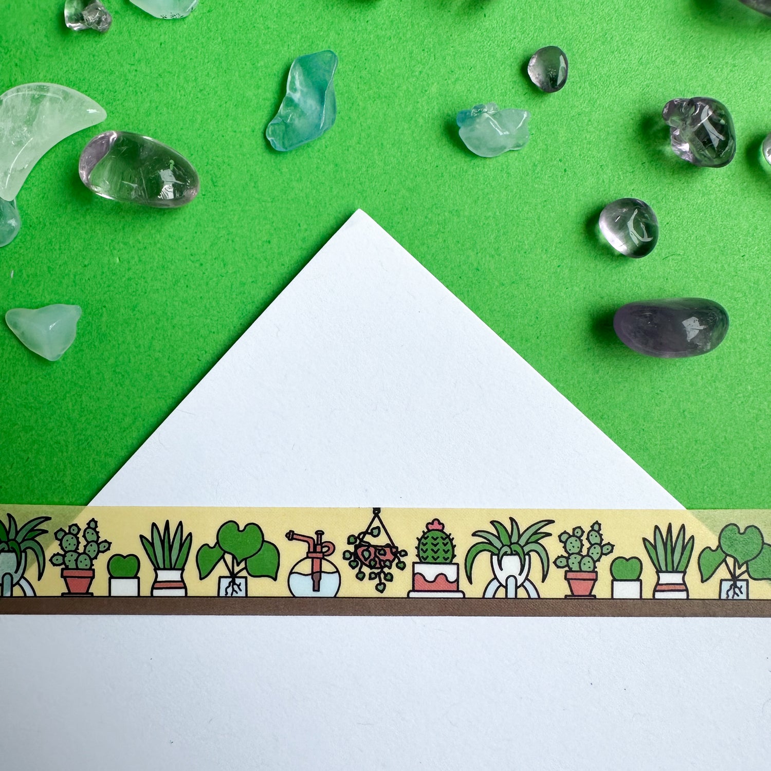 A corner of a white piece of paper with washi tape stuck to it with doodles of cute house plants on it. The paper is on a green piece of paper with crystals strewn around it. 