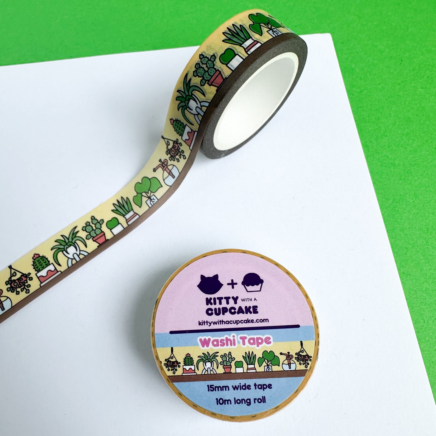 A roll of washi tape stuck to a white piece of paper, the design on the tape is different cute illustrations of house plants on a shelf. There is a packaged roll of the tape below.